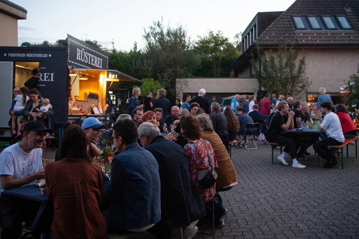 Menschen sitzen in der Dämmerung auf Festbänken, essen und trinken.